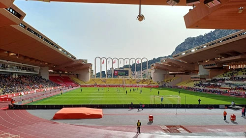 Blick aus der Fankurve auf das Spielfeld des Stade Louis II in Monaco während eines Spieltags. Im Hintergrund sind die charakteristischen Bögen und die felsige Landschaft zu sehen.